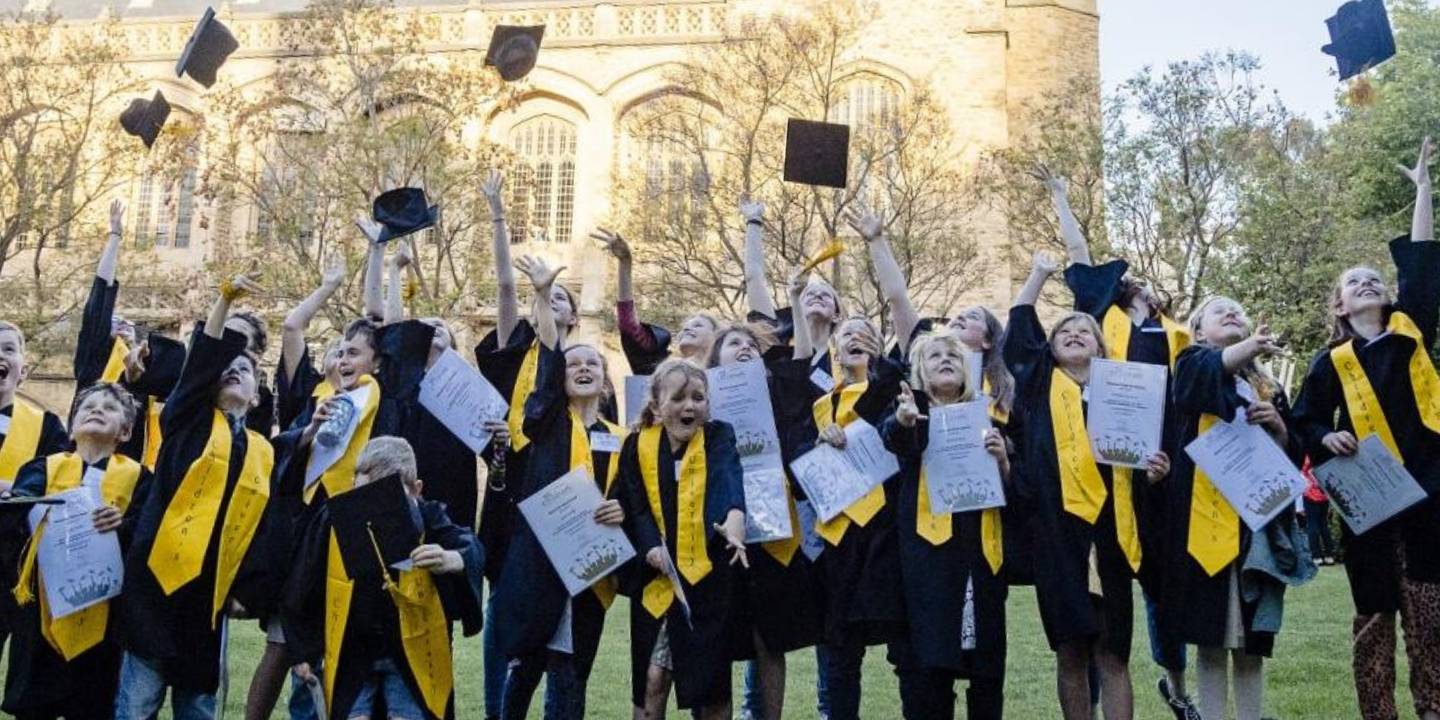 A big group of children in university graduation gowns throw their caps in the air in front of a university building.