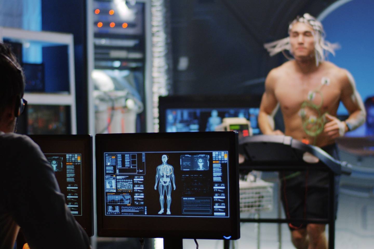 A man at a computer looks on at anatomical information that is being relayed via motion sensors on an athlete on a treadmill who is blurred in the background.
