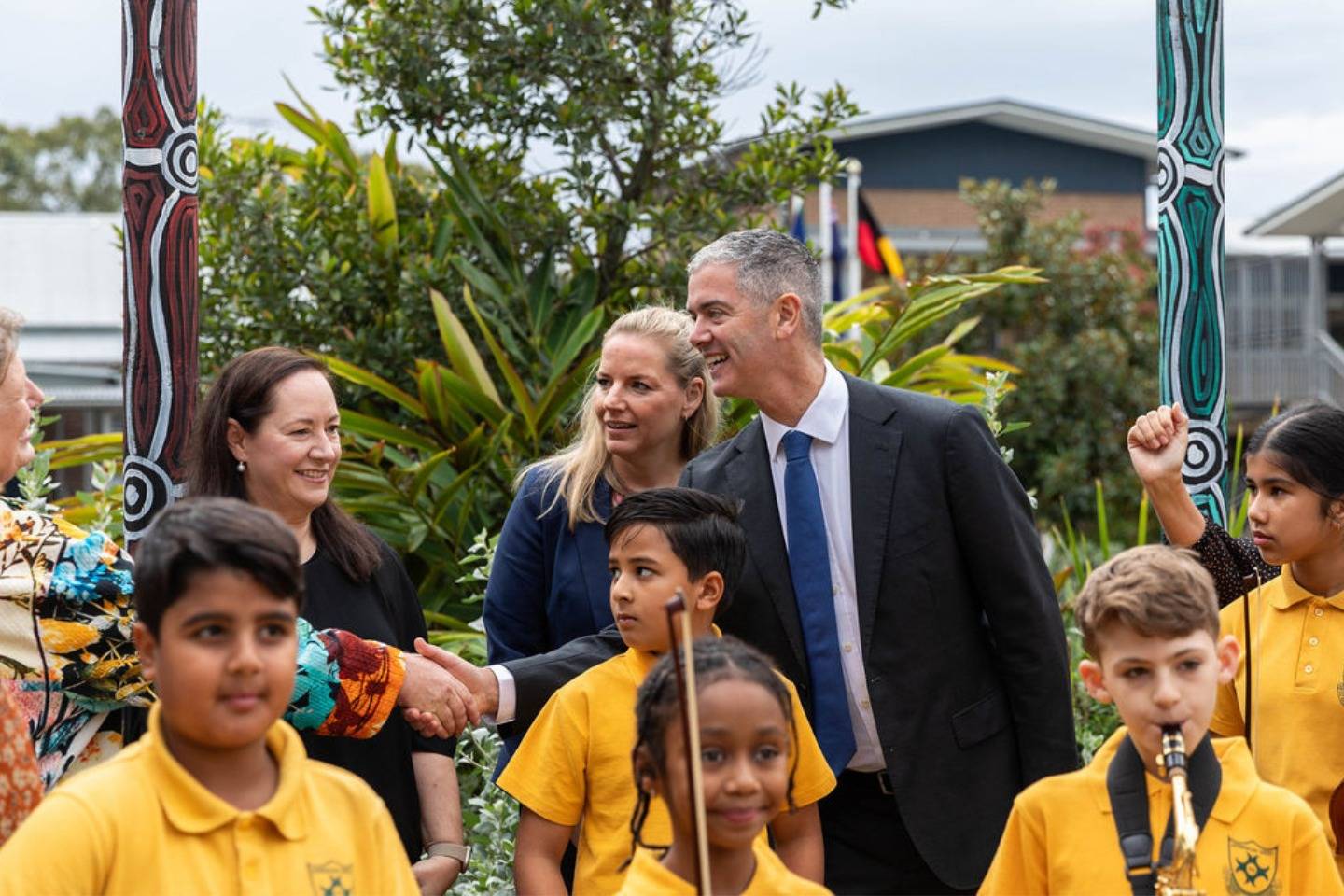 The NSW Minister for the Arts, Music and the Night-time Economy John Graham leans in front of  Dr Rebecca Taylor and Dr Anita Collins to shake the hand of a woman.  There are 5 primary students in the foreground in yellow tops carrying musical instruments.