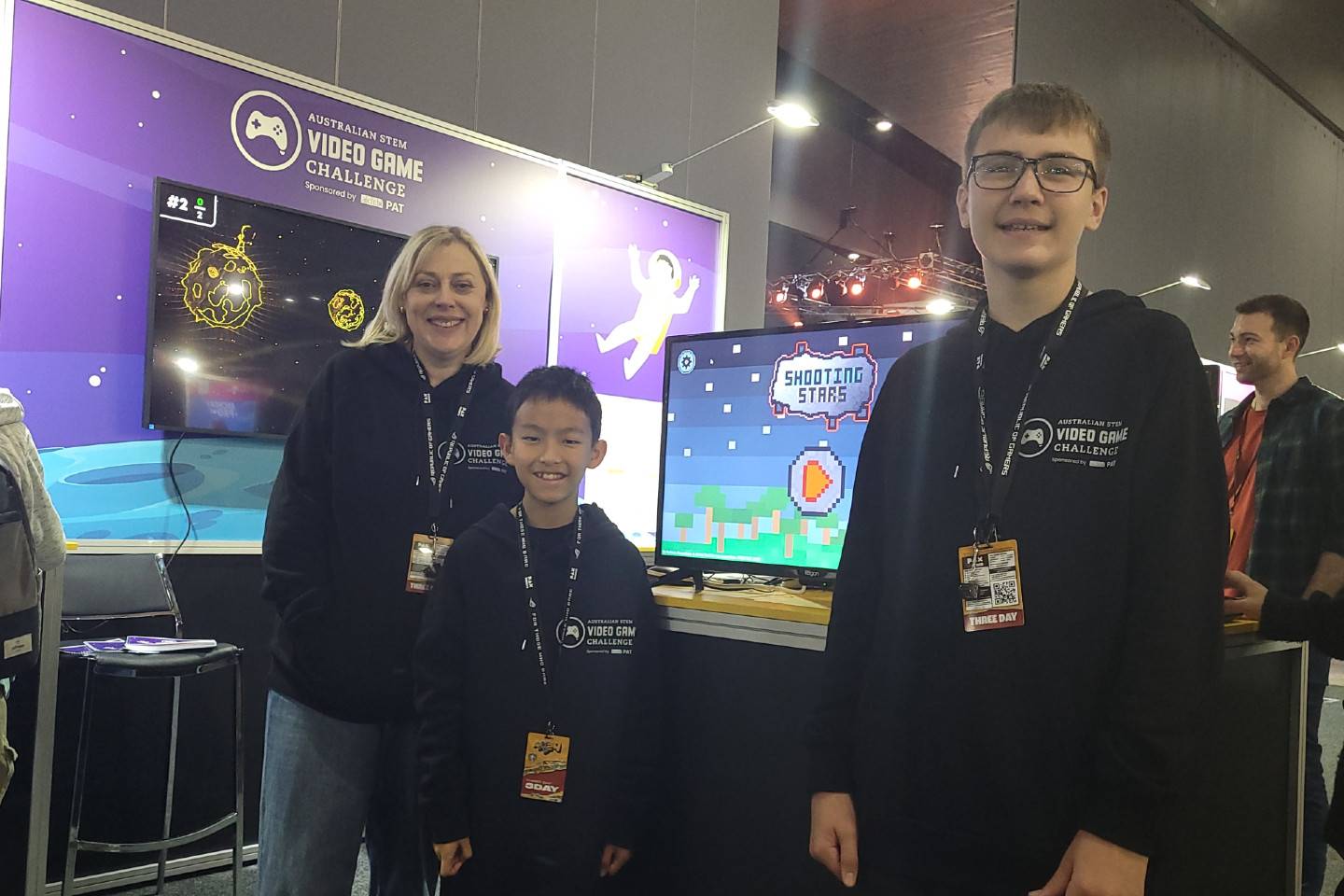 Two winning students and their challenge mentor in front of the Australian STEM Video Game Challenge stand at PAXAus gaming expo. Their game, 'Shooting Stars' is visible on a monitor.