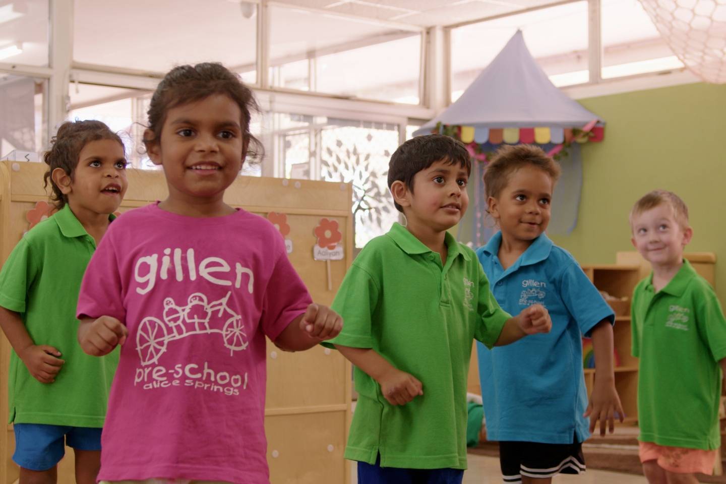 Five young children in colourful t-shirts are smiling and dancing in a preschool setting.
