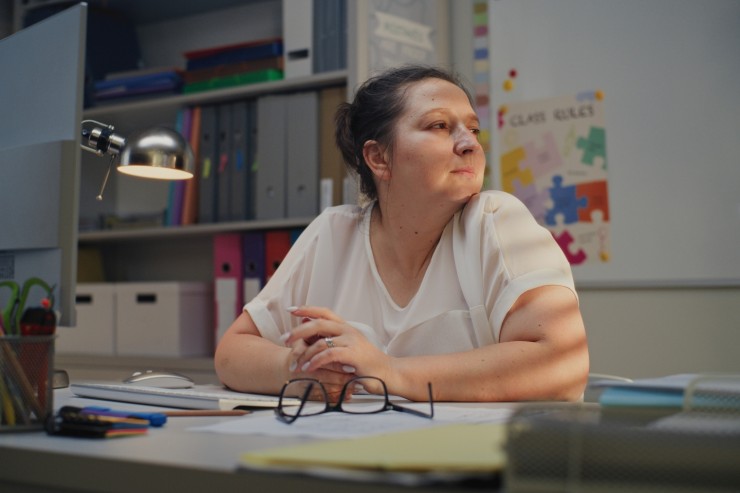 A female teacher sits at her desk with a lamp on, looking out a window.