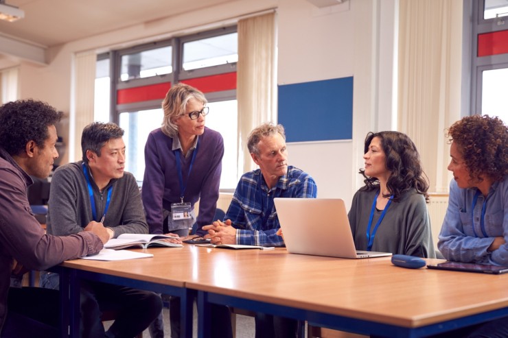 A group of educators sits around a table focusing on one female teacher with a laptop open who is leading a discussion.  Another teacher is standing at the end of the desk looking at the discussion leader.