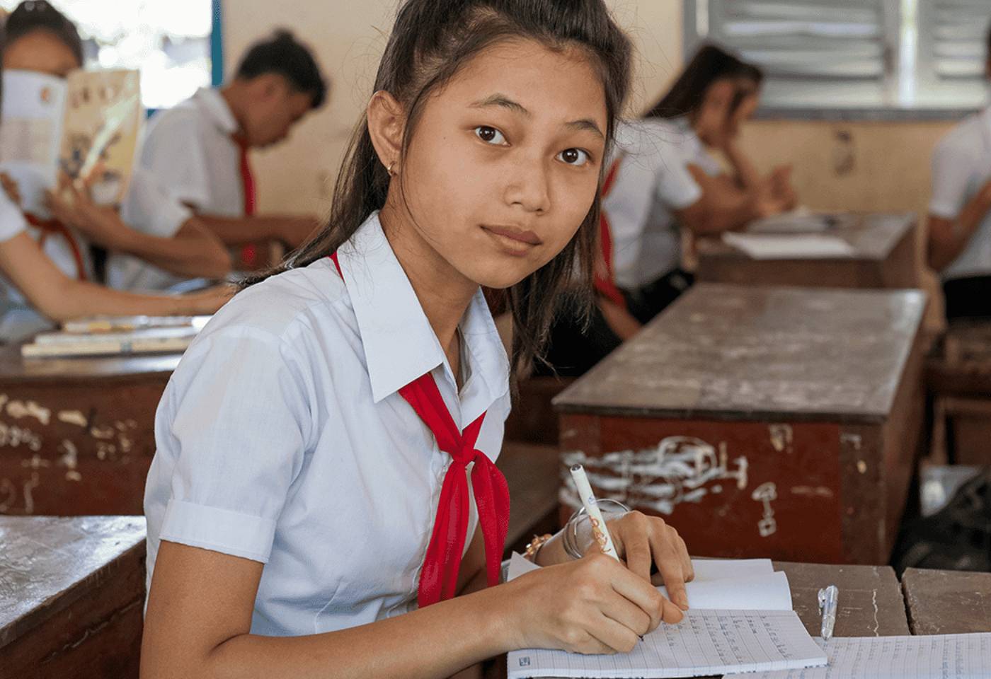 Stock image of a Vietnamese teenaged school girl wearing school uniform of a white short-sleeved shirt and a red scarf. She is writing with a pen on an exercise book, seated at a wooden desk. There are other students at desks in the background .
