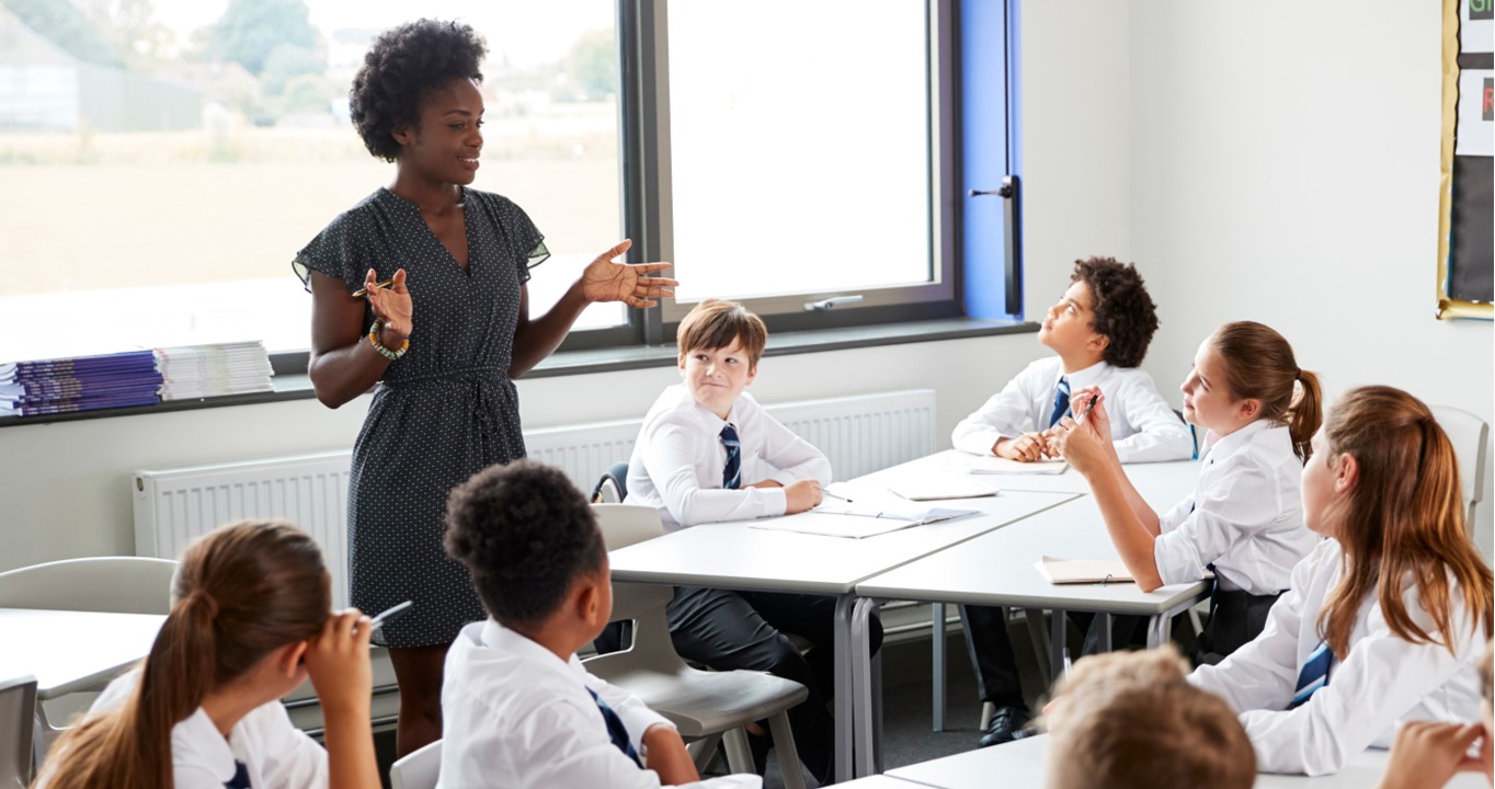 Stock image of a female teacher in the classroom with students sitting at desks.