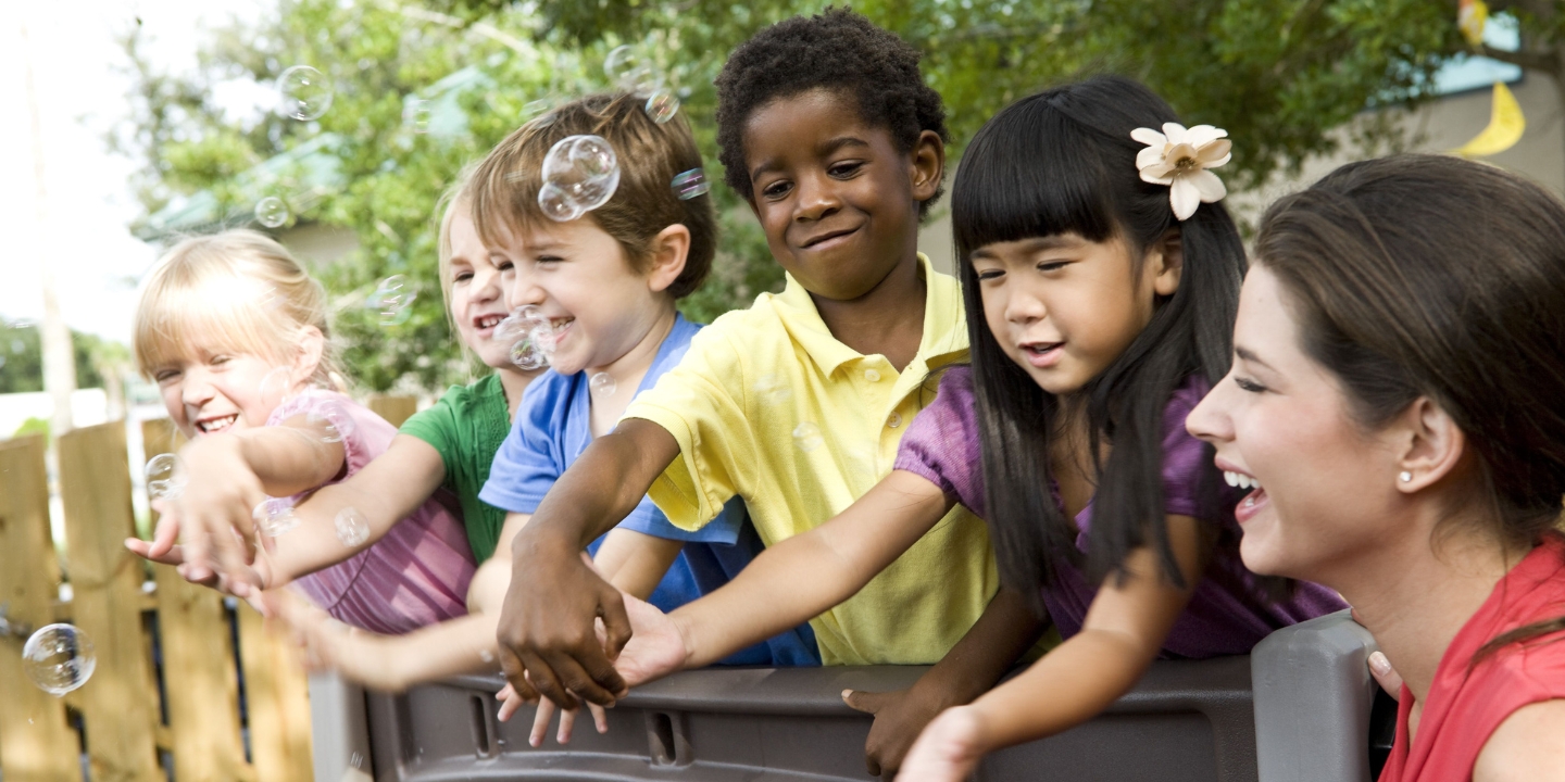 Young children lean over play equipment to bat away bubbles with teacher laughing beside them.