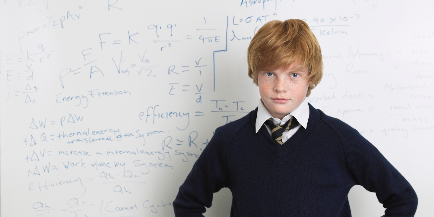 A teenage boy in school uniform stands in front of a whiteboard with maths equations on it, with a defiant look on his face.