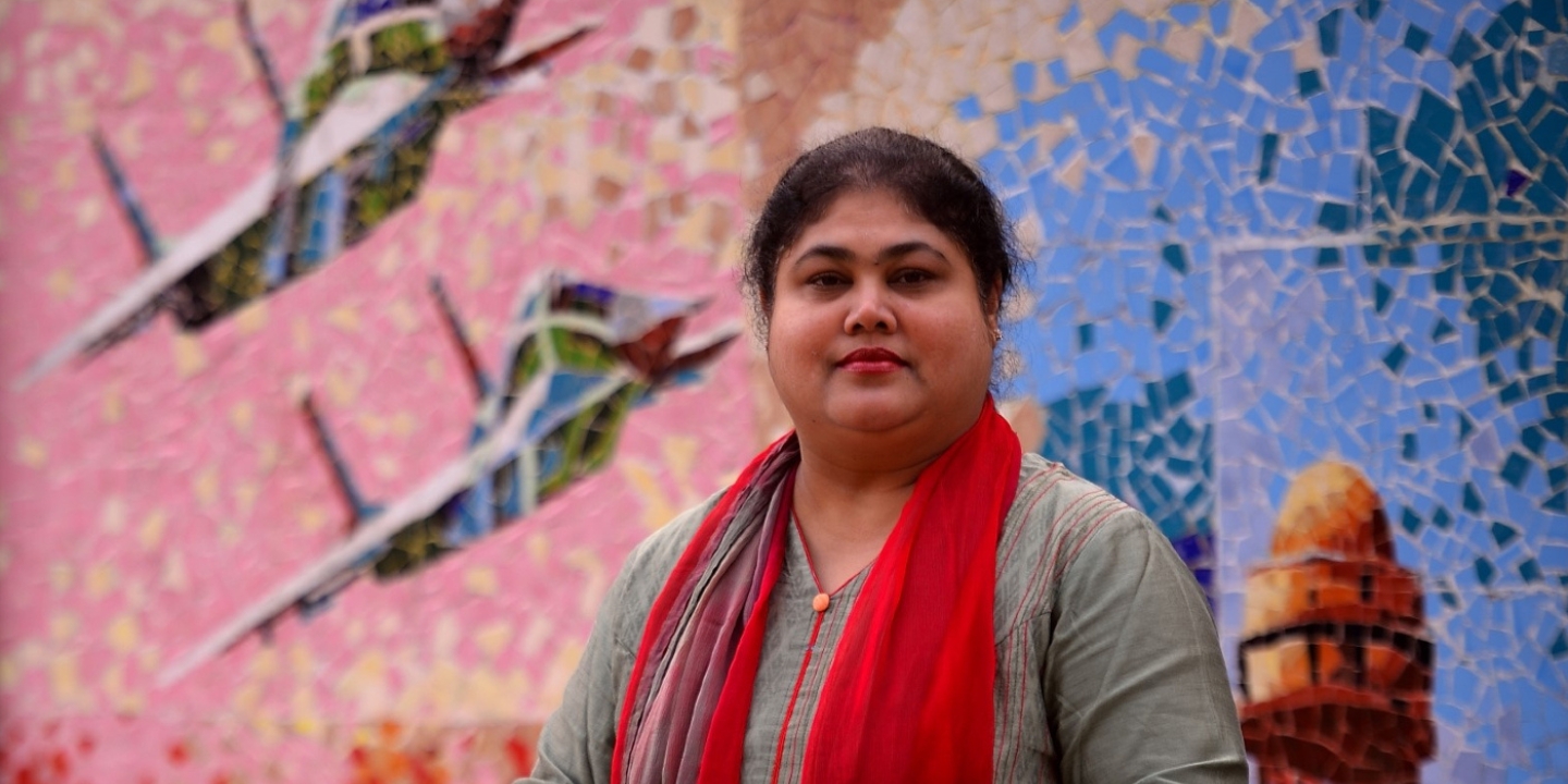 A woman with a red scarf draped around her neck stands in front of a colourful mosaic featuring two planes and a lighthouse. She is looking straight at the viewer, with only her head and shoulders in view.