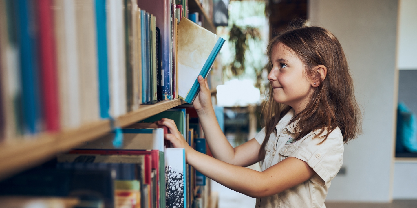 Female primary school student lifts a book out from a library shelf while smiling.