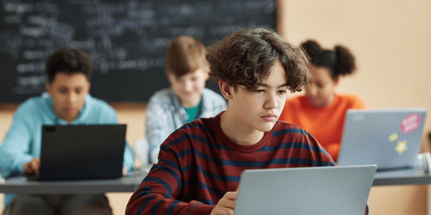 Stock image of teenager using a laptop in the foreground with other teenagers using laptops in the background.