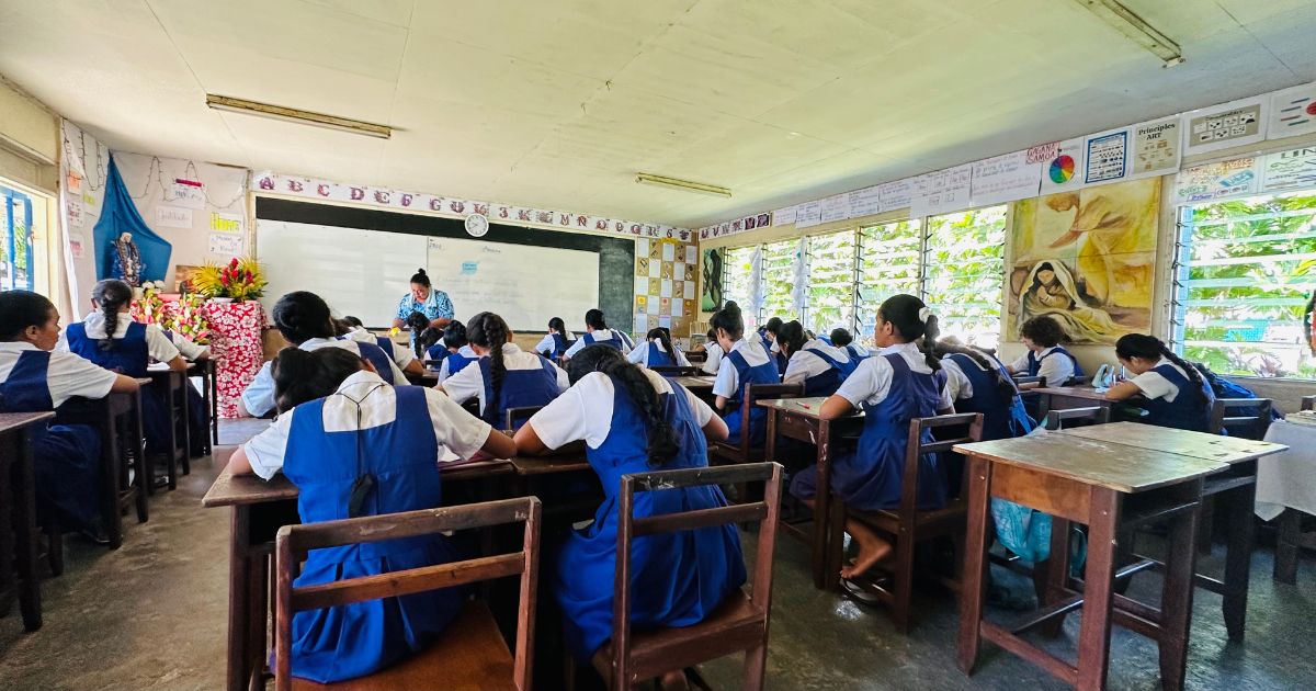 A stock image of children at their desks seen from the back of a Samoan classroom.