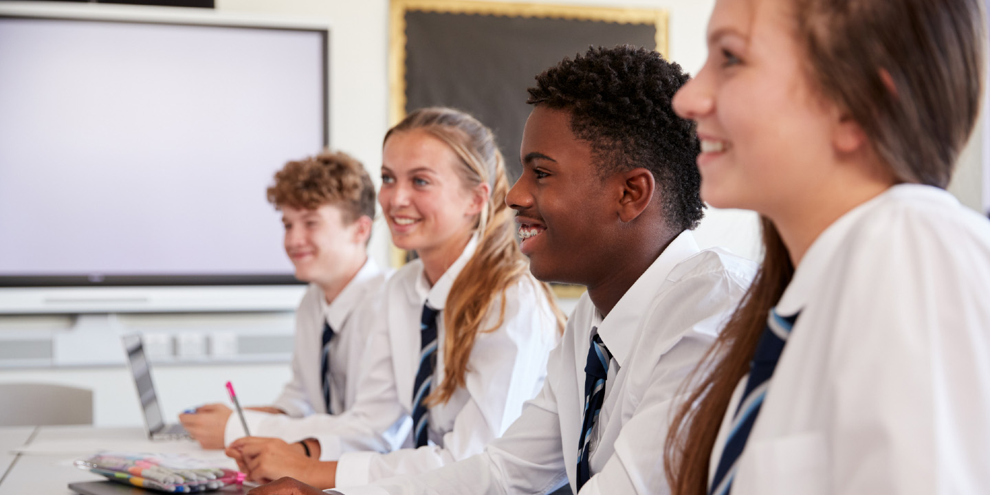 Line Of High School Students Wearing Uniform Sitting At Desk In Classroom