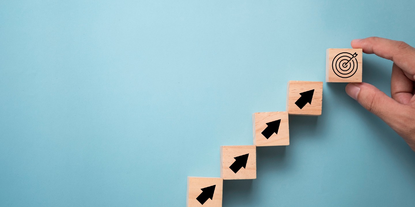 Stock image of hand stacking a wooden block with an archery target printed on it, in a diagonal line with wooden blocks with arrows pointing north-east, indicating an upward trend.