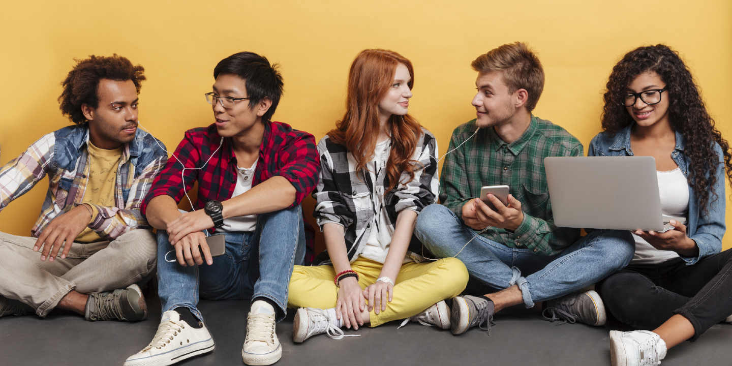 Stock image of multiethnic group of cheerful young people sitting on floor listening to music and using laptop over yellow background.