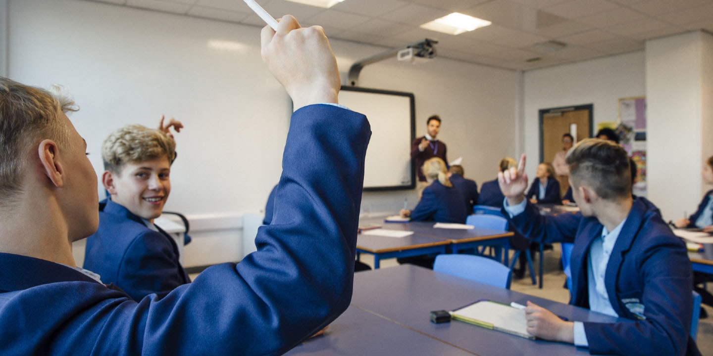 Point of view shot of a high school lesson where the teacher has asked a question and some students have their hands up to answer.