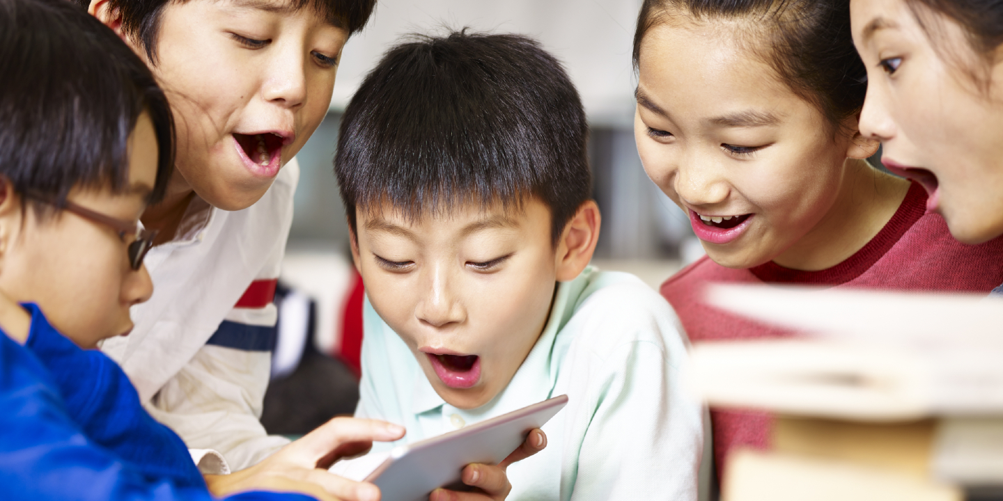 Stock image of group of asian elementary school children gathering around playing game together using tablet device.
