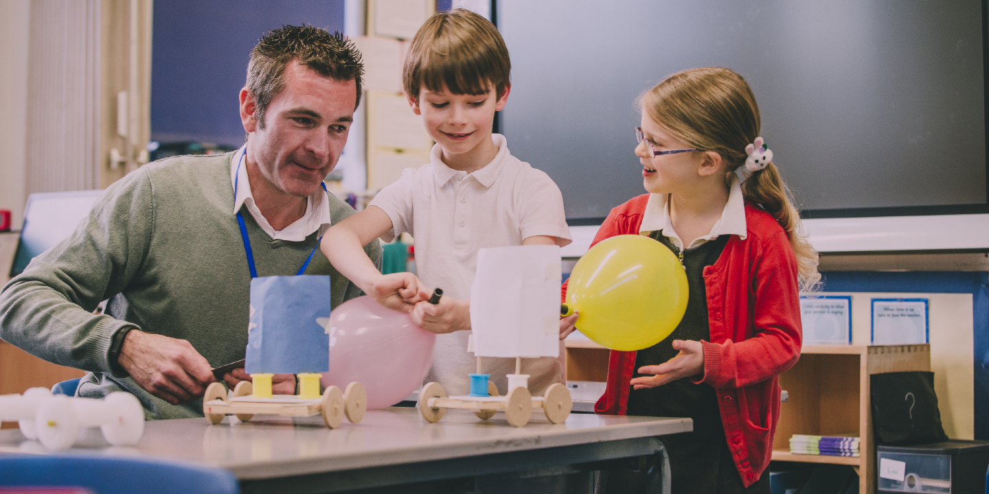 Stock image of a primary school teacher helping two of his students build wind-powered cars using recycled items and crafts equipment.