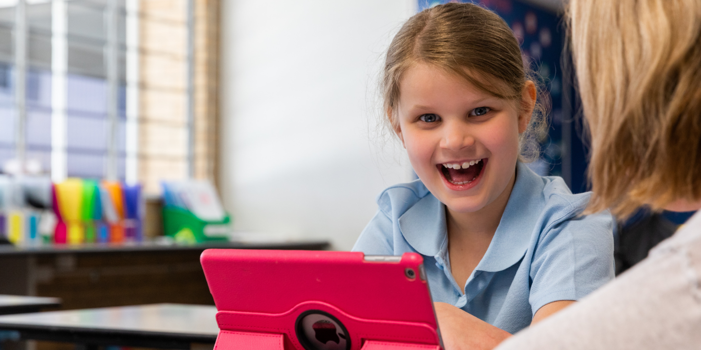 A young schoolgirl sitting with her red iPad is overseen by her teacher. The student is smiling.