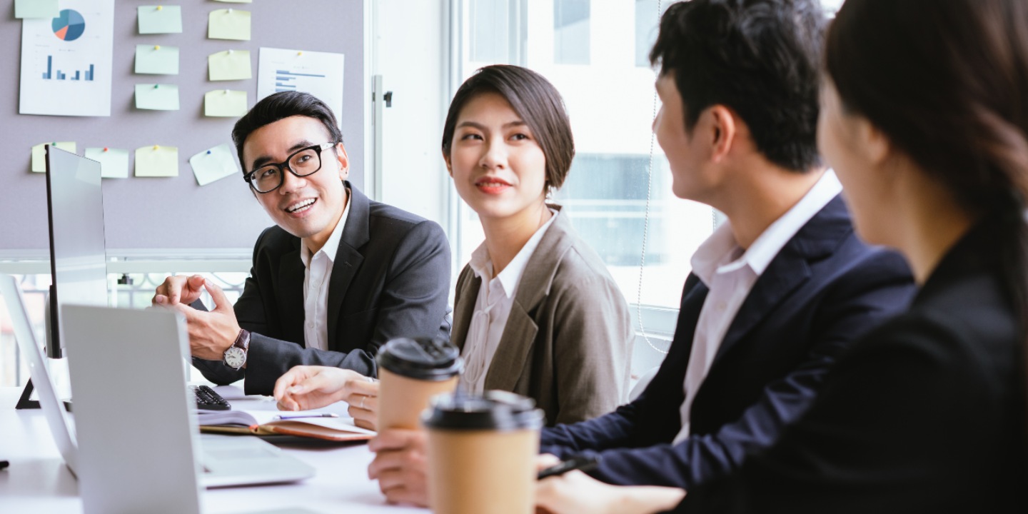 The photo shows a group of male and female business people wearing corporate clothes who are sitting in a row at a desk with open laptop computers and coffees in disposable coffee cups