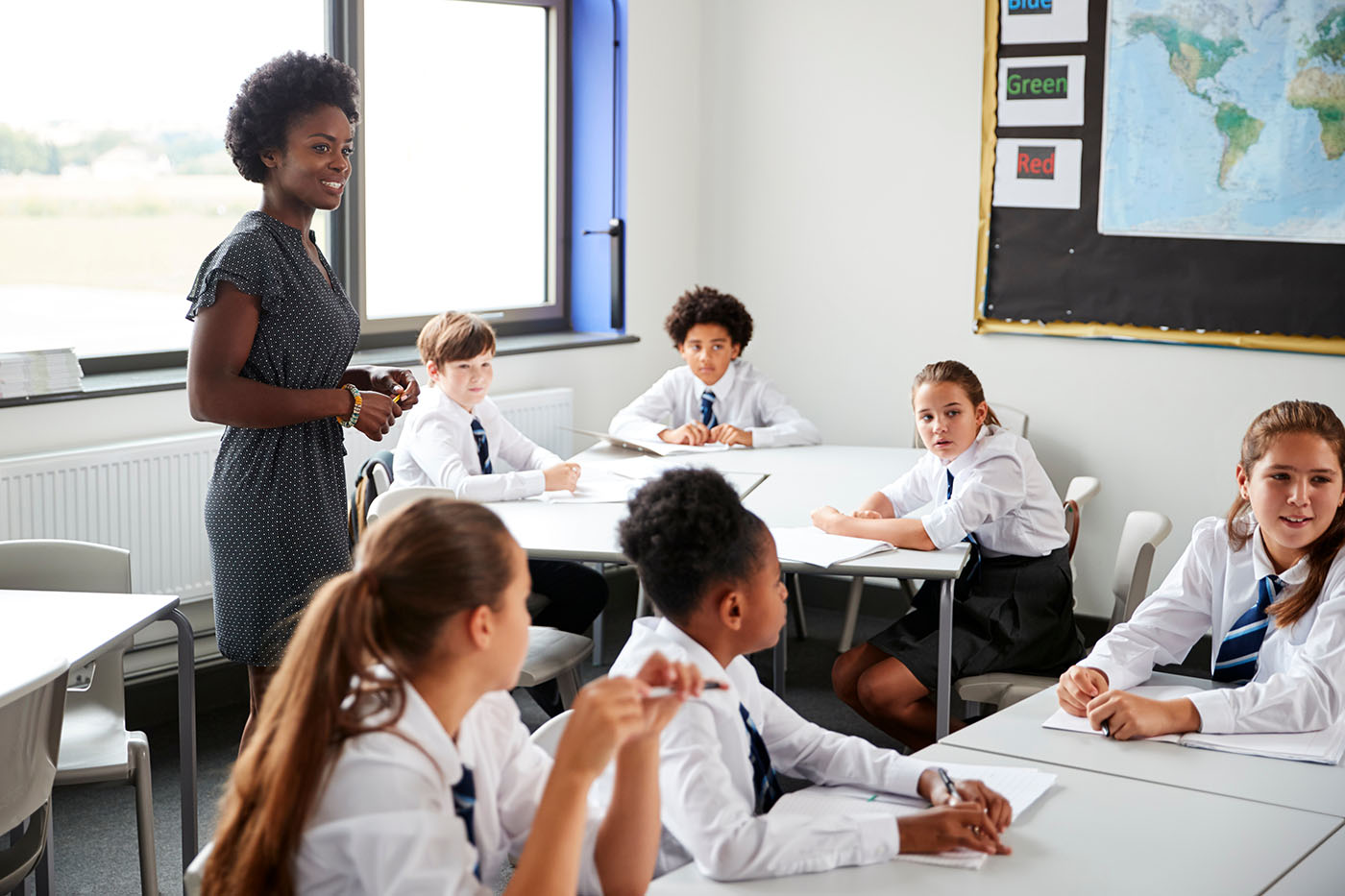 Female High School Tutor Helping Students Wearing Uniform Seated Around Tables In Lesson