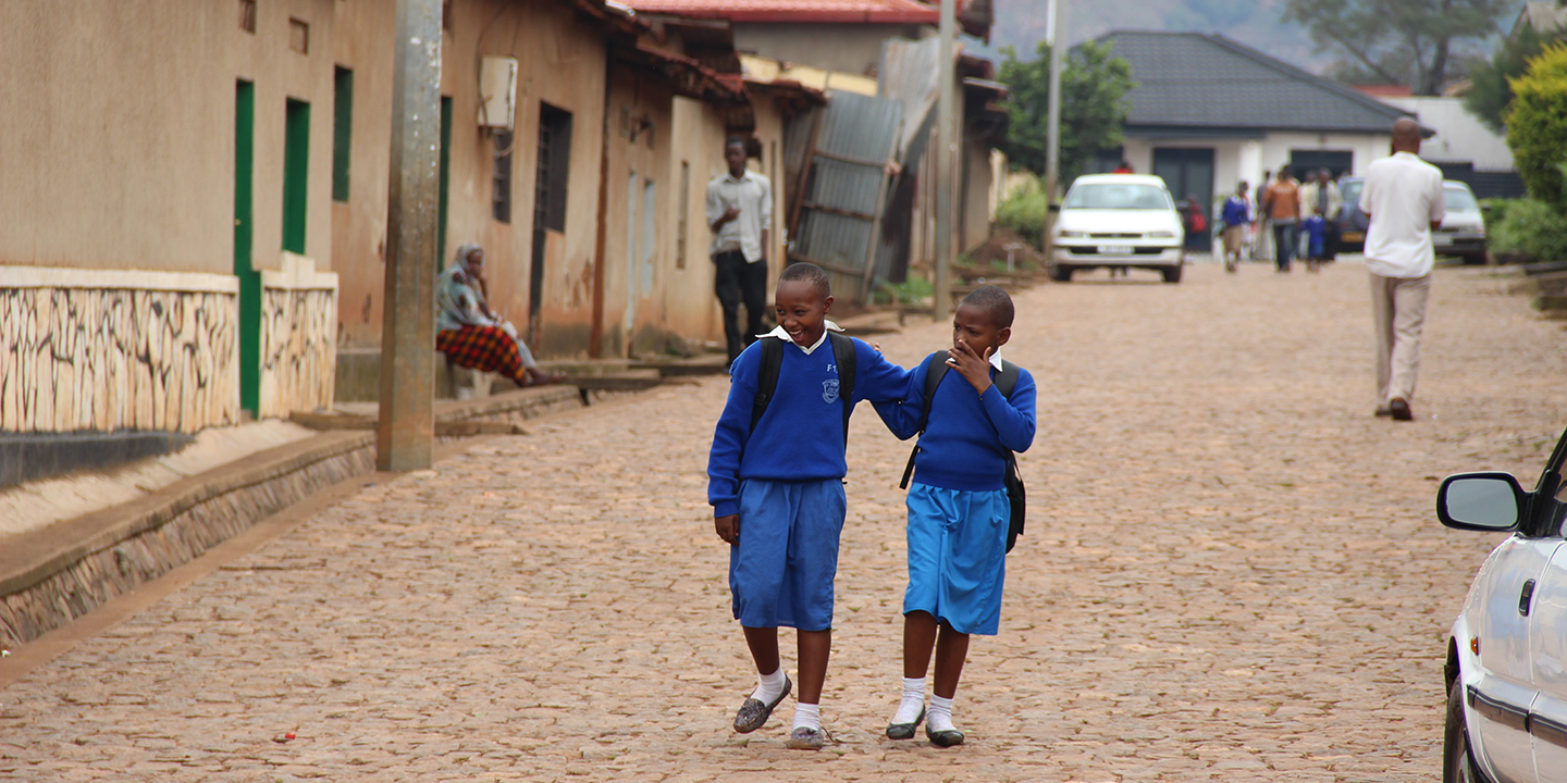 Two school children in blue uniforms walking through a village with backpacks on and arms around each other