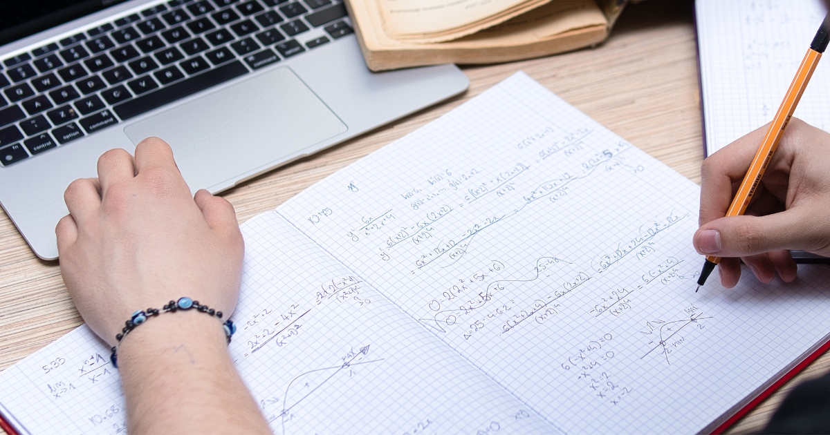 Stock image of a teenager's hands. One is holding a pencil and working on maths equations in a workbook and the other is resting on an open laptop.