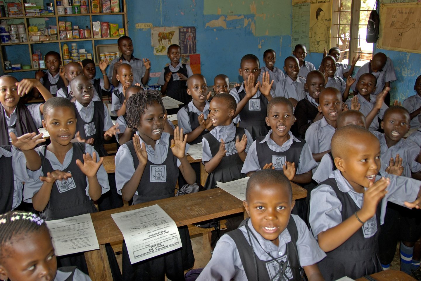 Children in uniform are smiling and clapping their hands at their desks in a Ugandan school.