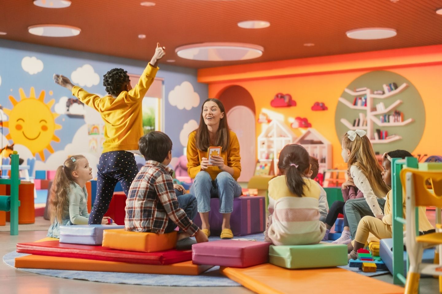 A female educator among a circle of sitting children in a cheerily decorated kindergarten smiles as a young girl jumps up.