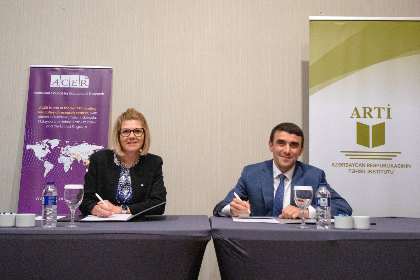 ACER CEO Lisa Rodgers and Institute Director Elnur Alieyev are seated alongside each other and signing a document each with banners from their organisations behind them.