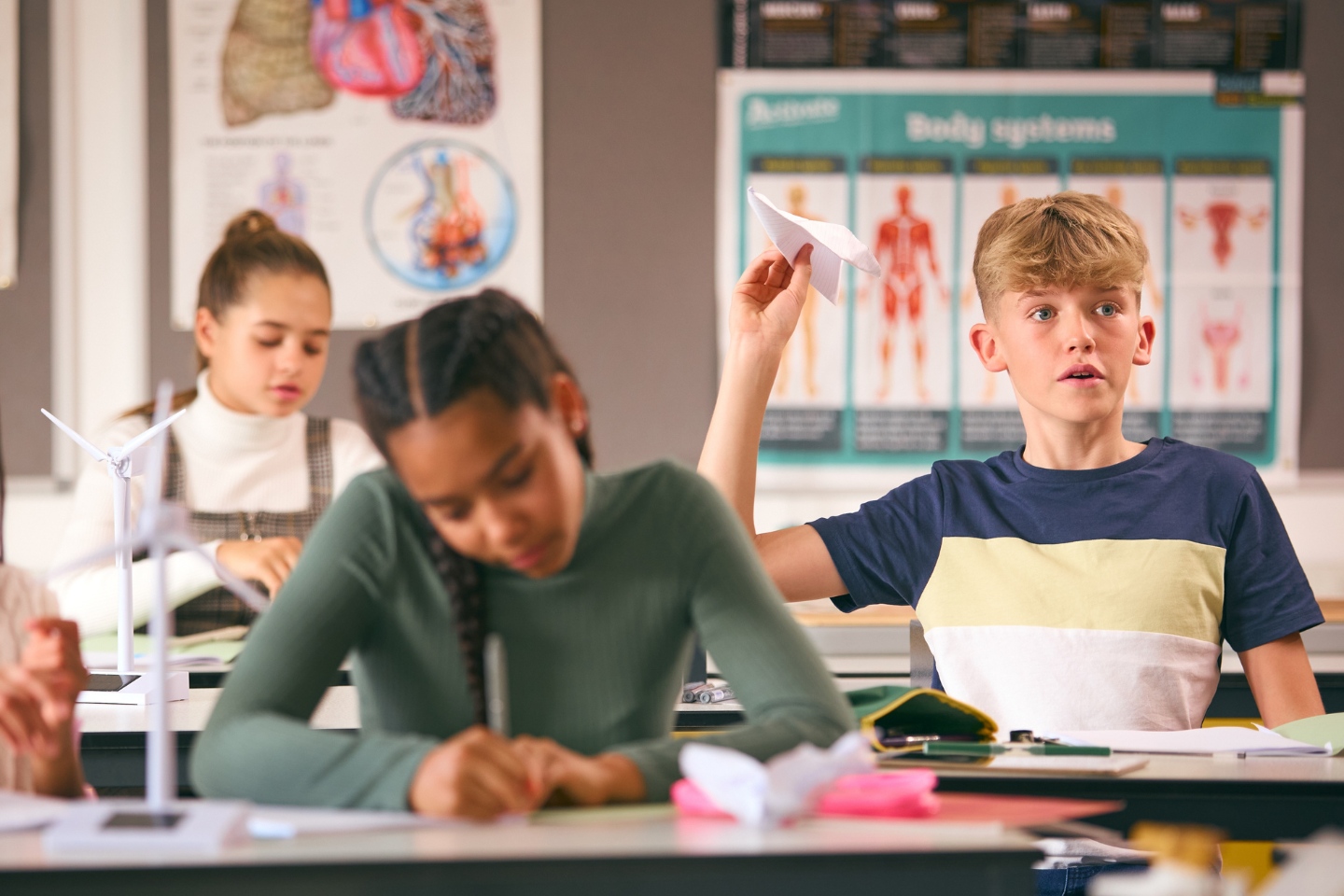 A boy sits in class with a paper plane held aloft ready to throw. Two girls are seated near him but are blurred.