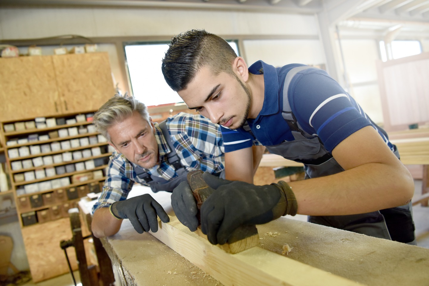 A young man in gloves works with wood as a male teacher leans over a workbench to check the work.